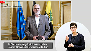 Das Bild zeigt einen Ausschnitt aus dem Grußwort von Rainer Robra, Staatsminister und Minister für Kultur des Landes Sachsen-Anhalt. Im Hintergrund sind drei Flaggen zu erkennen, unter anderem die Landesflagge und die EU-Flagge. Am unteren Bildrand sind die Untertitel zu sehen. Am rechten Bildrand befindet sich die Dolmetscherin für Gebärdensprache.
