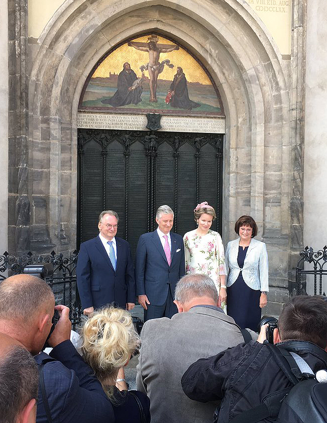 Das Foto zeigt das belgische Königspaar, flankiert von Ministerpräsident Haseloff und seiner Gattin, vor der Thesentür der Schlosskirche Wittenberg. Im Vordergrund sind mehrere Fotografen zu sehen.