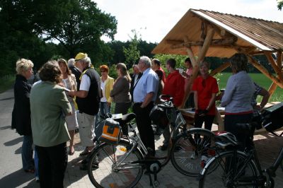 Mehrere Personen mit Fahrrädern stehen auf dem Radweg vor dem Haltepunkt mit überdachtem Tisch und Bänken aus Holz.