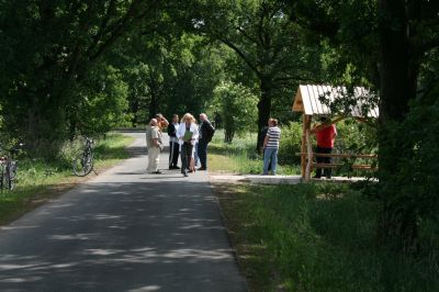 Asphalt-Radweg zwischen Bäumen. Auf der rechten Seite ein Haltepunkt mit überdachtem Tisch und Bänken aus Holz. Davor stehen mehrere Personen.
