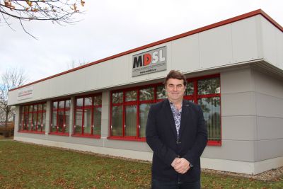 Auf dem Foto sieht man ein weißes Gebäude mit roten Fensterrahmen und einem Schild mit der Aufschrift "MDSL". Vor dem Gebäude steht der Geschäftsführer. (Foto: Ministerium der Finanzen LSA)