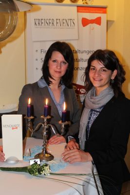 Das Bild zeigt zwei junge Frauen an einem Stehtisch auf dem ein dreiarmiger Kerzenständer mit brennenden Kerzen steht.