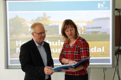 The picture shows Prof. Michael Hoffmann and Ilona Darius standing in front of a projection screen, giving a presentation. Both are looking at a brochure Ilona Darius holds in his hand. (Picture: Ministry of Finance of Saxony-Anhalt)
