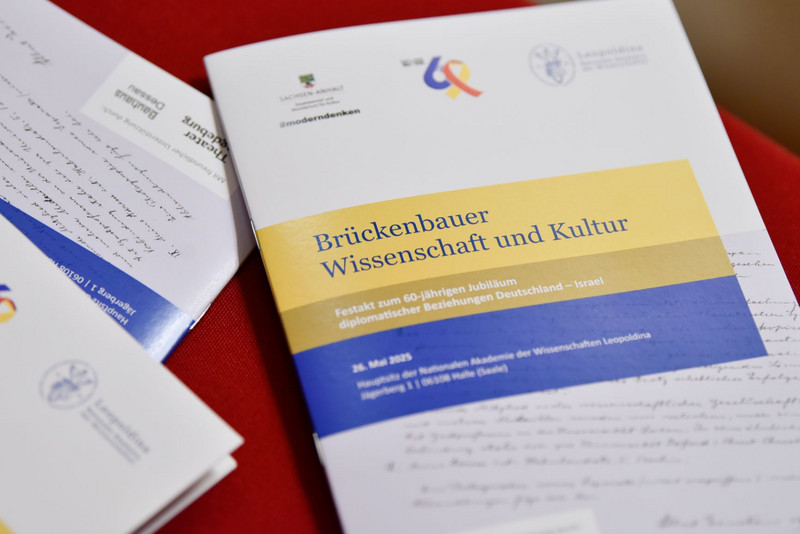 Das Programmheft des Festakts liegt auf einem Tisch. Auf dem Cover sind oben Logos, in der Mitte steht der Titel "Brückenbauer Wissenschaft und Kultur", geschrieben in blau auf einem gelben Hintergrund.