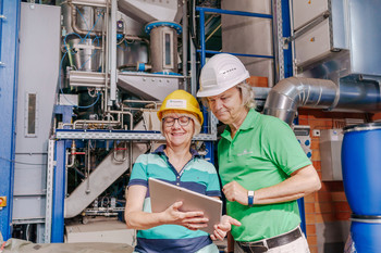 Das Bild zeigt zwei Personen mit einem Tablett vor einer Industrieanlage. 