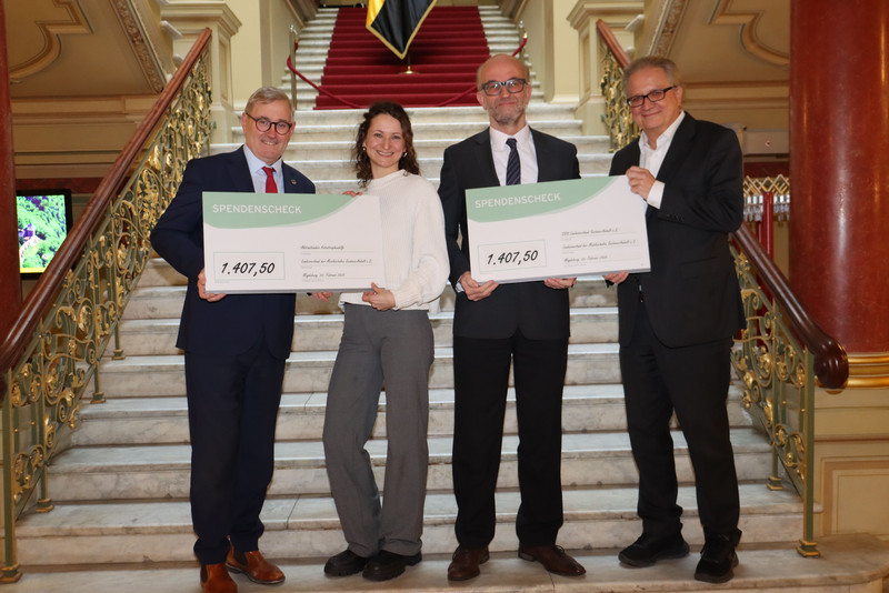 Im Treppenhaus der Staatskanzlei stehen vier Menschen. Sie tragen jeweils einen Spendenscheck von 1407,50 euro. Links steht Staatssekretär Dr. Jürgen Ude, Denise Landmann, Senior Managerin Fundraising Aktionsbündnis Katastrophenhilfe, Dr. Christian Reineke, Geschäftsführer des Landesverbands der Musikschulen Sachsen-Anhalt, Dr. Carlhans Uhle, Geschäftsführer Landesverband DRK