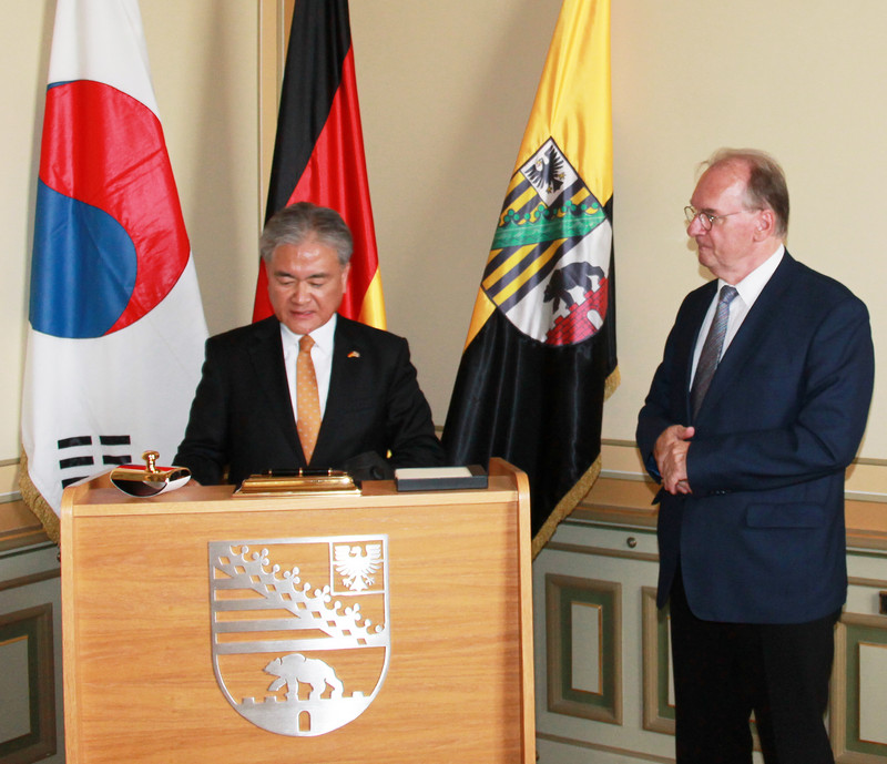 Das Bild zeigt Botschafter Jong hinter einem mit dem Landeswappen versehenen Pult bei der Eintragung in das Gästebuch der Landesregierung. Rechts schaut Ministerpräsident Haseloff zu. Im Hintergrund stehen die Flaggen Südkoreas, Deutschlands und Sachsen-Anhalts.