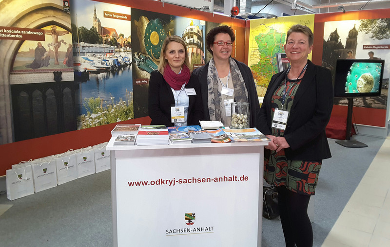Das Bild zeigt hinter einem Tresen mit dem Landeslogo Ministerin Prof. Dr. Claudia Dalbert (r.) am Sachsen-Anhalt-Stand auf der World Travel Show mit den Standbetreuerinnen Agnieszka Misiuk und Angelika Beulecke.