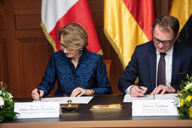 Das Bild zeigt links die französische Botschafterin Descôtes und rechts Bildungsminister Tullner, an einem Tisch vor den Flaggen Deutschlands, Frankreichs und Sachsen-Anhalt sitzend, beim Handschlag nach der Unterzeichnung der Vereinbarung zur Zusammenarbeit im Bildungsbereich.