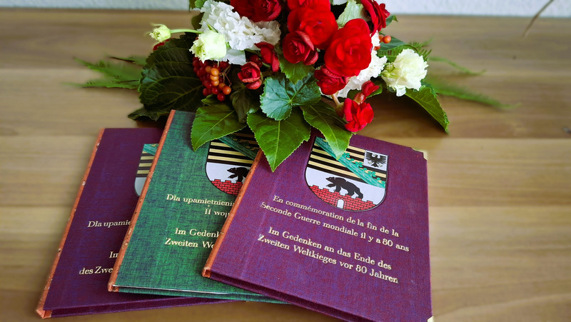 Drei Bücher, zwei in bordeauxrot und eins in grün, liegen auf einem Tisch. Im Hintergrund steht ein Blumengesteckt mit roten und weißen Blumen. Auf dem Cover der Bücher steht "Im Gedenken an das Ende des Zweiten Weltkrieges vor 80 Jahren" auf Deutsch und in der jeweiligen Sprache.