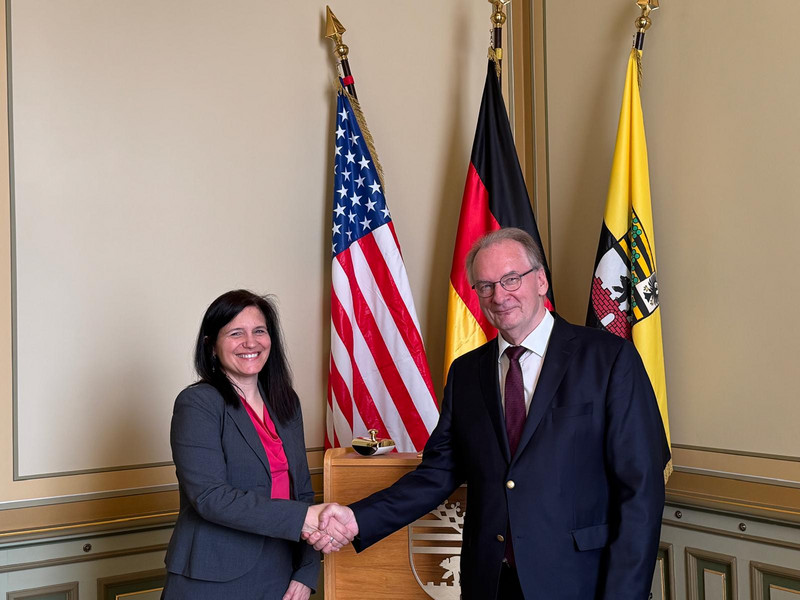 Die Generalkonsulin steht links. Sie und der Ministerpräsident geben sich die Hand und schauen in die Kamera. Im Hintergrund sind die Flaggen der USA, Deutschland und Sachsen-Anhalt zu sehen. 