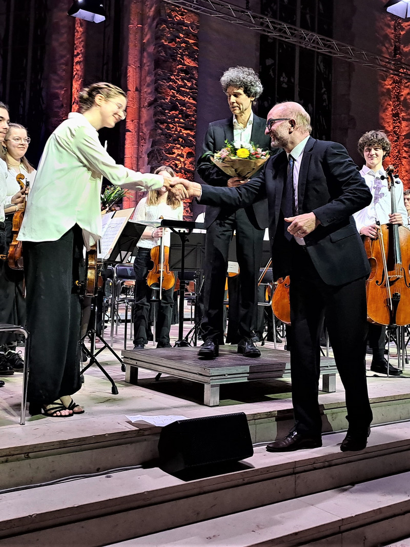 Links steht eine junge Frau. In der Mitte steht der Dirigent und hält einen Blumenstrauß in den Händen. Rechts steht Dr. Christian Reinecke und schüttelt die Musikerin die Hand. 