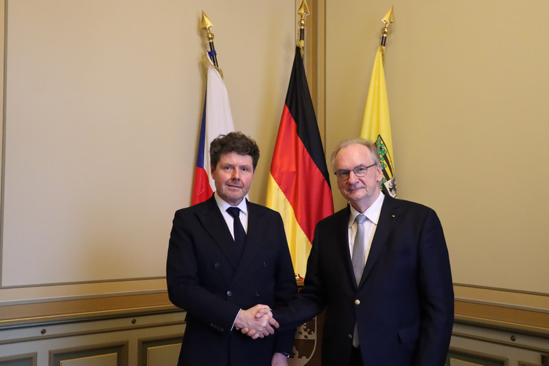 Beide Männer tragen Anzüge und geben sich die Hände. Im Hintergrund sind die Flaggen der Tschechischen Republik, Deutschland und Sachsen-Anhalt von links nach rechts zu sehen.