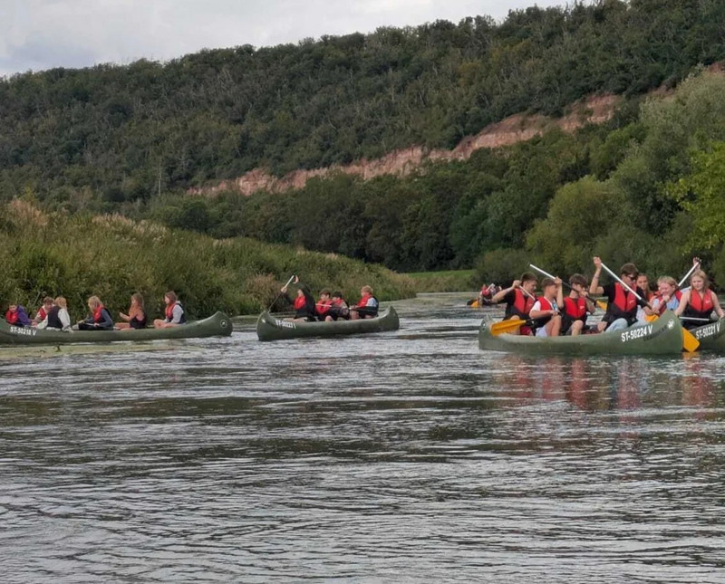 Es sind vier Kanus auf dem Wasser, in jedem sitzen vier Jugendlichen. Im Hintergrund ist ein bewaldeter Hang zu sehen.