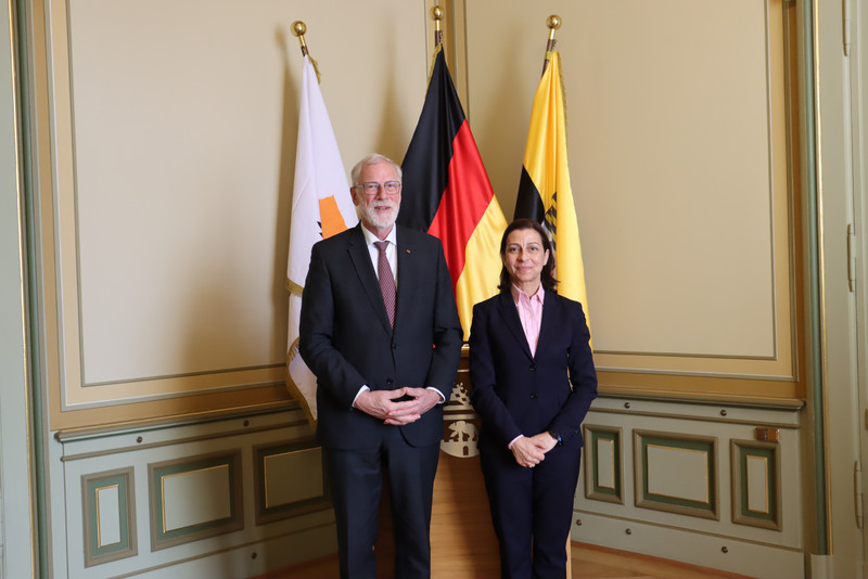 Beide tragen schwarze Anzüge und stehen nebeneinander. Im Hintergrund sind drei Flaggen: von links nach rechts Zypern, Deutschland und Sachsen-Anhalt