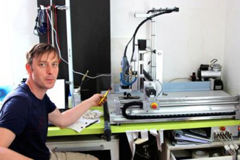 Das Foto zeigt den Geschäftsführer und Entwickler David Goll im blauen T-Shirt. Er sitzt an einem Tisch mit einer Fräsmaschine; mitten im Labor Labor der Firma Lagotec. (Foto: Friedemann Kahl)