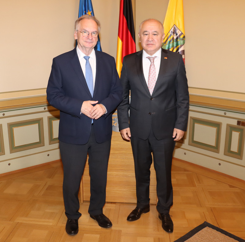 Ministerpräsident Reiner Haseloff steht links und hält sich die Hände. Der Botschafter steht daneben. Im Hintergrund sind drei Fahnen zu sehen.