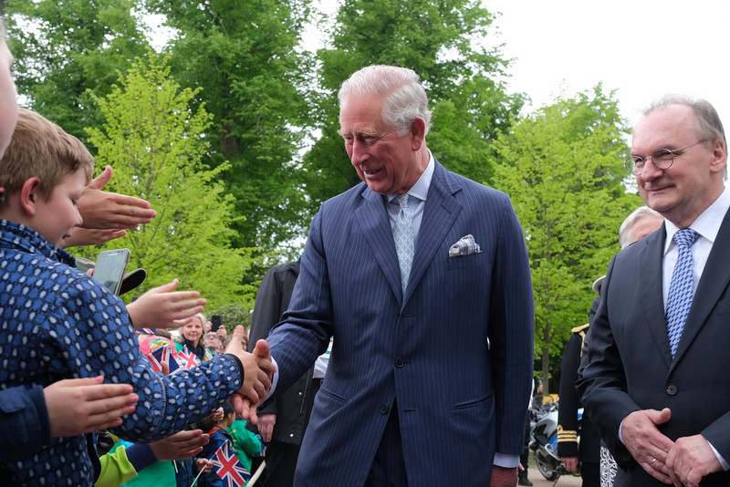 Das Bild zeigt in der Mitte Prinz Charles, der einem Jungen, der mit vielen anderen Kindern und Erwachsenen zur Begrüßung des Gastes vor dem Schloss wartete, die Hand schüttelt. Rechts im Bild ist Ministerpräsident Haseloff zu sehen.