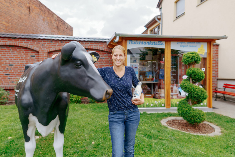 Junglandwirtin Andrea Finke vor Ihrer Milchtankstelle an eine Kuh-Statue gelehnt