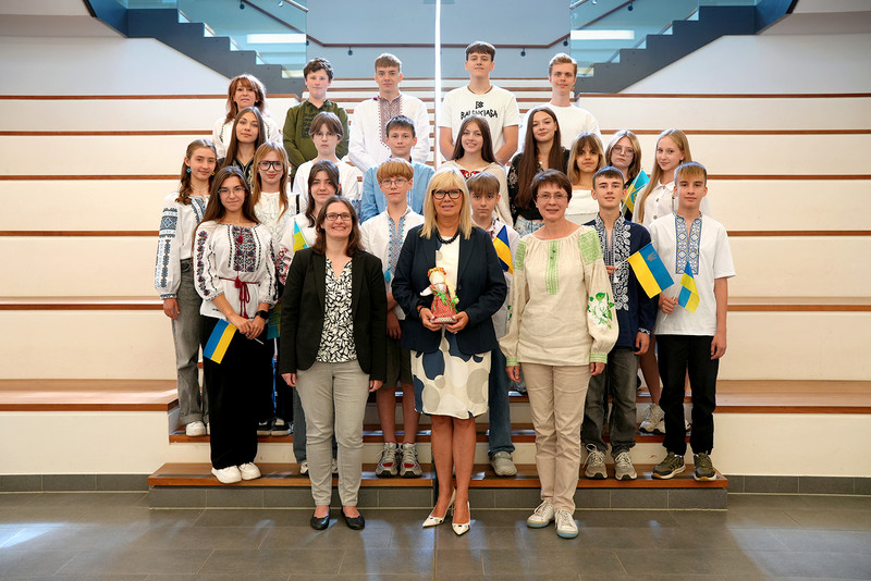Eine Gruppe Kinder steht auf einer Treppe. Vorne unten steht Oberbürgermeister Borris und hält in den Hände eine Motanka-Puppe.