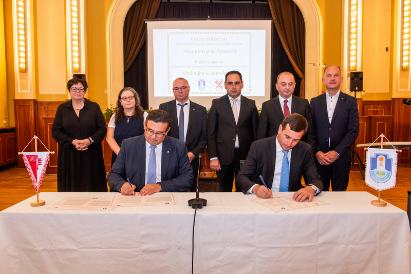 Zwei Männer sitzen an einem Tisch und unterschreiben ein Dokument. Im Hintergrund stehen zwei Frauen (links) und vier Männer. 
