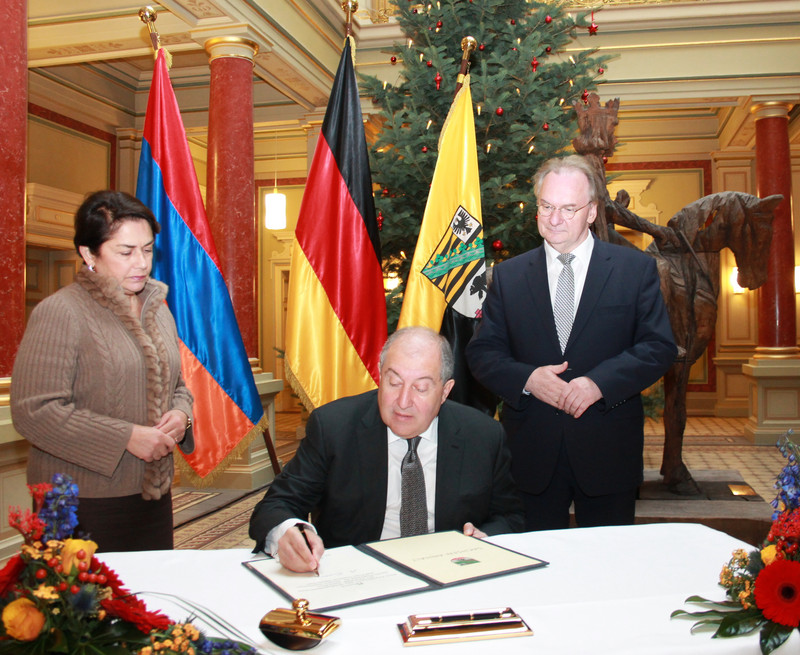 Das Bild zeigt an einem Tisch sitzend den Präsidenten Armeniens bei der Eintragung ins Gästebuch des Landes Sachsen-Anhalt. Links neben ihm steht sein Gattin und rechts Ministerpräsident Reiner Haseloff. Hinter den Personen stehen die Flaggen Armeniens