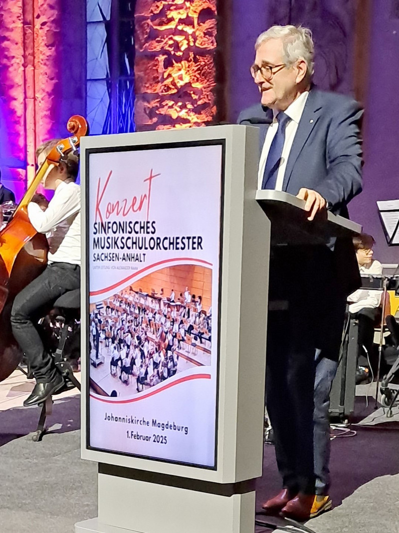 Staatsekretär Jürgen Ude trägt einen Anzug und steht an einem grauen Pult. Vorne auf dem Pult ist das Plakat der Veranstaltung zu sehen. Links sitzt ein Musiker mit einer Kontrabass. 