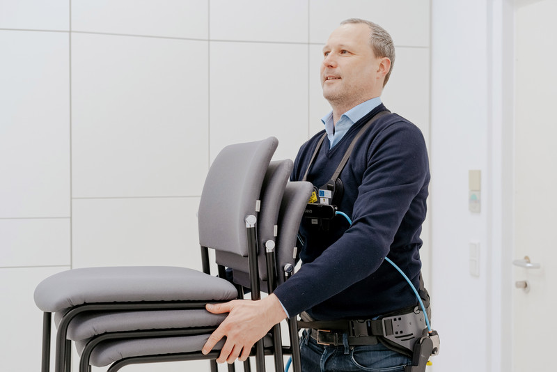 Das Bild zeigt eine Mann mit einem Exoskelett, der Stühle trägt.  