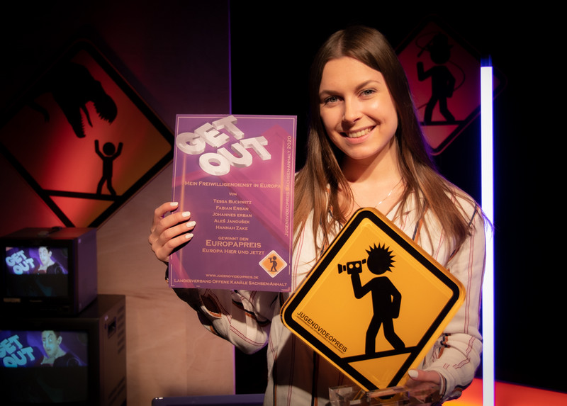 Das Bild zeigt Tessa Buchwitz als Vertreterin des Gewinnerteams in der Sonderkategorie „Europa – hier und jetzt“ einen Pokal mit Jugendvideopreis-Emblem und eine lilafarbene Urkunde haltend.