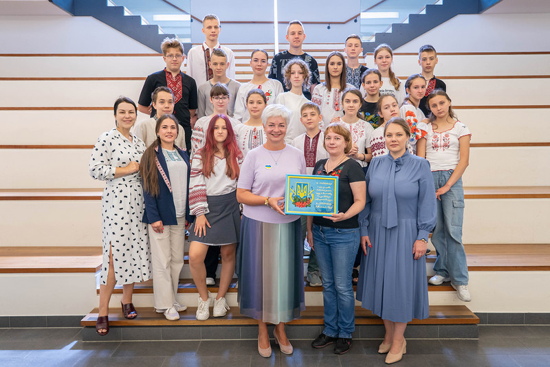Eine Gruppe mit etwa 20 Kinder steht auf einer Treppe. Vorne in der Mitte steht die Bürgermeisterin und sie hält in den Händen ein Bild mit einem Logo und Text auf ukrainisch in blau-gelb.