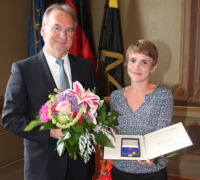 Das Bild zeigt links Ministerpräsident Dr. Reiner Haseloff, der Violaine Varin (rechts) die Ehrennadel des Landes Sachsen-Anhalt und einen Blumenstrauß überreicht.