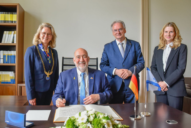 Der Botschafter Ron Proso sitzt an einem Tisch vor einem Gästebuch. Links daneben steht Prof. Dr. Bettina Rockenbach, Präsidentin der Leopoldina, rechts des Botschafters stehen Ministerpräsdient Dr. Reiner Haseloff und Franziska Hornig, Generalsekretärin der Leopoldina.