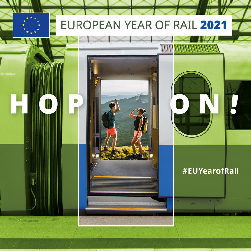 Das Bild zeigt im Hintergrund in grünem Farbton den Abstand eines im Bahnhof stehenden Zuges. Durch die geöffnete Eingangstür blickt der Betrachter auf einen vollfarbigen Bildausschnitt mit einem jungen Pärchen, das sich vor einer Bergkulisse mit einer High-Five begrüßt. Im Vordergrund steht der weiße Schriftzug "Hop on!" sowie rechts unten der Hashtag "EU Year of Rail".