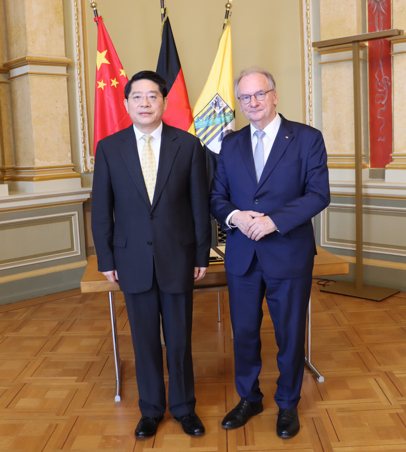 Beide Personen stehen nebeneinander. Botschafter Hongbo Deng trägt einen schwarzen Anzug, Ministerpräsident Haseloff einen dunkelblauen. Im Hintergrund sind drei Flaggen zu sehen: links die Flagge der Volksrepublik China, in der Mitte Deutschland und rechts Sachsen-Anhalt.