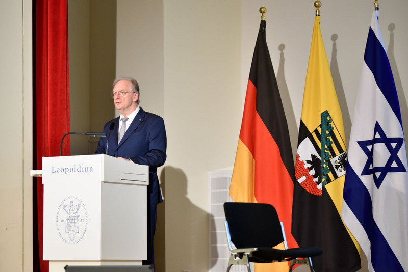 Links steht Ministerpräsident Haseloff an einem weißen Pult. Darauf steht vorne Leopoldina und deren Logo. Rechts stehen drei großen Fahnen, von links nach rechts Deutschland, Sachsen-Anhalt und Israel