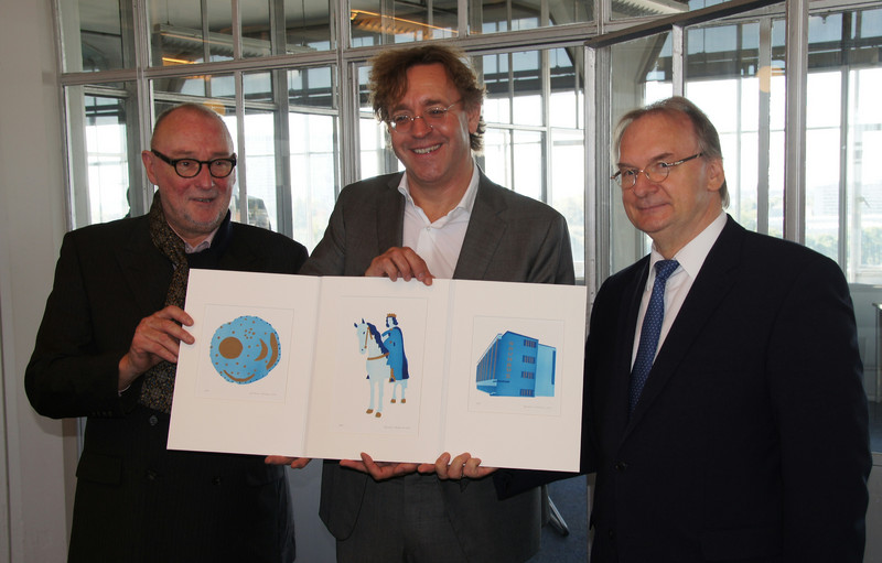 Das Bild zeigt von links Gastgeber Leonard Kooy und Roger Maertens sowie Ministerpräsident Reiner Haseloff mit einer von ihm überreichten dreiteiligen Grafikmappe aus Sachsen-Anhalt, darunter eine Ansicht des Bauhauses Dessau.