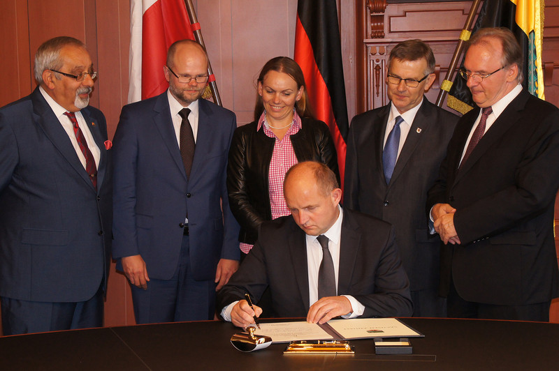 Das Foto zeigt Marschall Całbecki, der sich an einem Tisch sitzend in das Gästebuch der Staatskanzlei einträgt. Dahinter stehen rechts Ministerpräsident Haseloff und die vier weiteren Delegationsmitglieder vor den Flaggen Polens, Deutschlands und Sachsen-Anhalts.