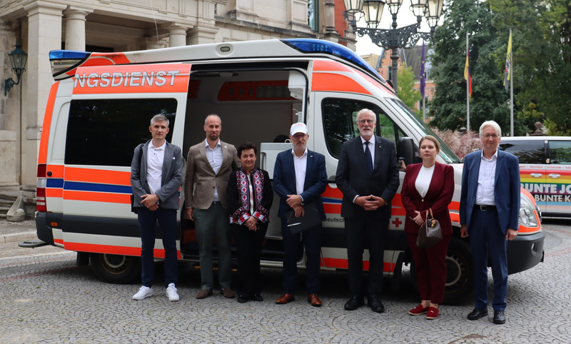 250917_Übergabe Krankenwagen Ukraine Ein Krankenwagen parkt mit offener Tür vor der Staatskanzlei. Davon stehen sieben Menschen in einer Reihe.