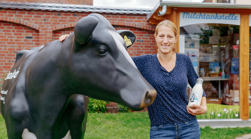 Das Bild zeigt Junglandwirtin Andrea Finke mit einer gläsernen Milchflasche im Arm, während sie die Hand auf den Nacken einer schwarzen Kunst-Kuh legt. Im Hintergrund ist das Schaufenster einer Milchtankstelle zu sehen.