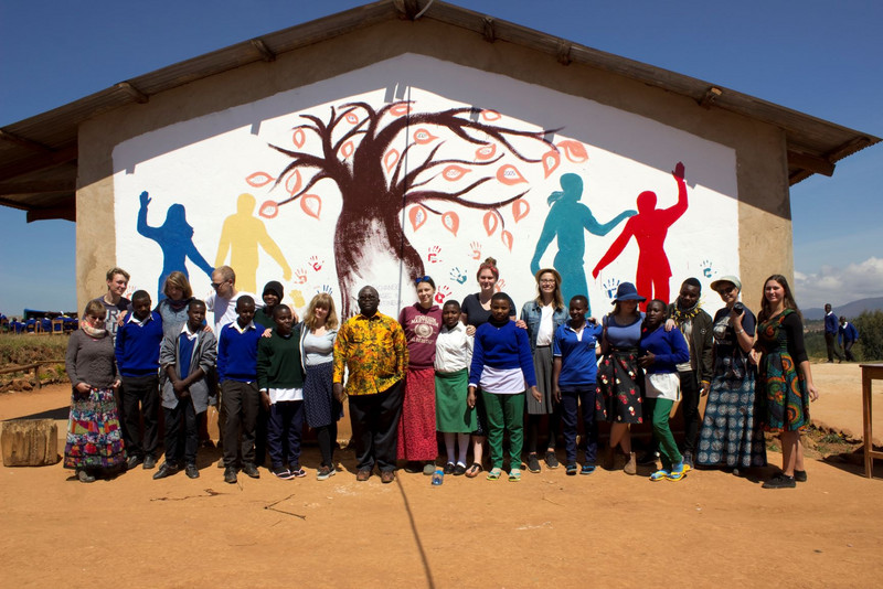 Das Bild zeigt eine Gruppe junger Menschen, die vor einer bunt bemalten Wand einer Schule in die Kamera lächeln.
